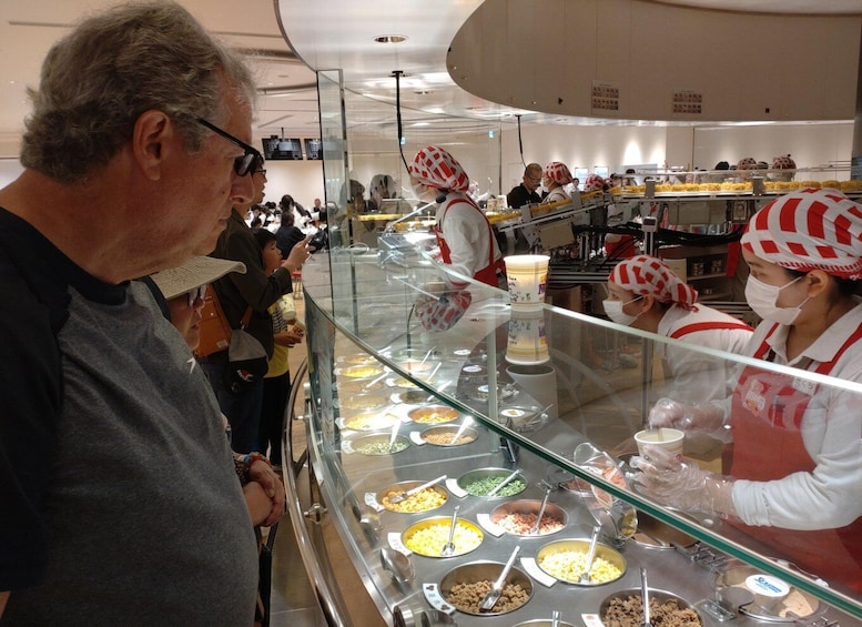 A buffet line with various food items in bowls, a man in the foreground, and staff members in the background wearing red and white uniforms. 1