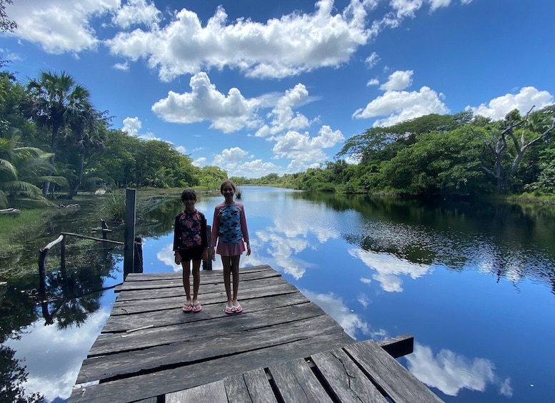 Two people standing on a wooden dock by a calm river surrounded by lush greenery. 4