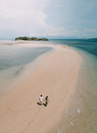A sandy beach with two people holding hands walking along the shore, a small island in the distance, and calm sea. 4