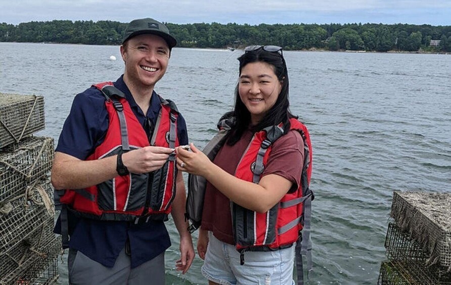 Two people wearing life jackets standing by a body of water with fishing traps and a forested shoreline in the background. 2