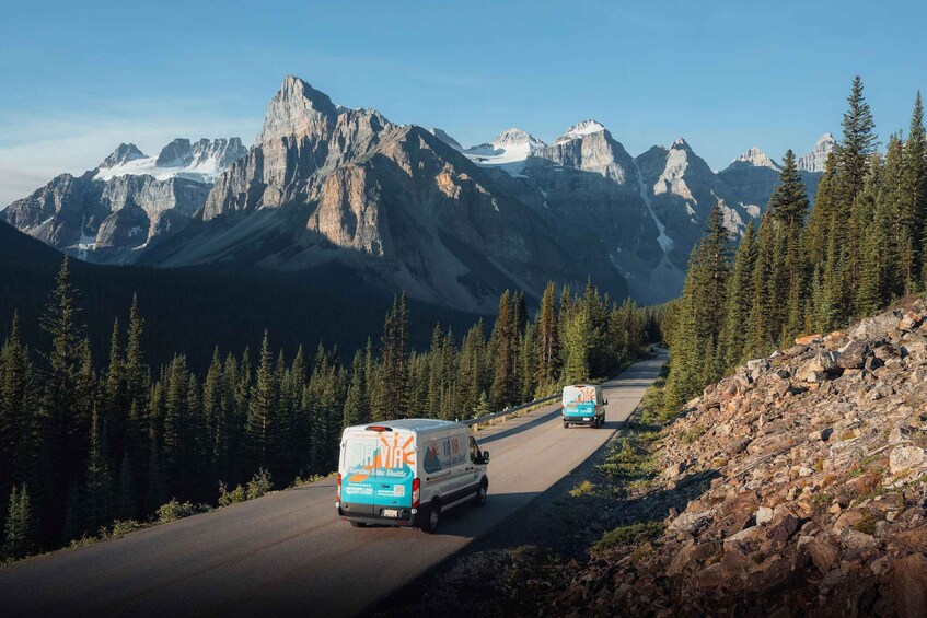 A road with two buses labeled 'VIA VIA' and 'VIA VIA' driving on a mountain road surrounded by dense forests and towering mountains. 4
