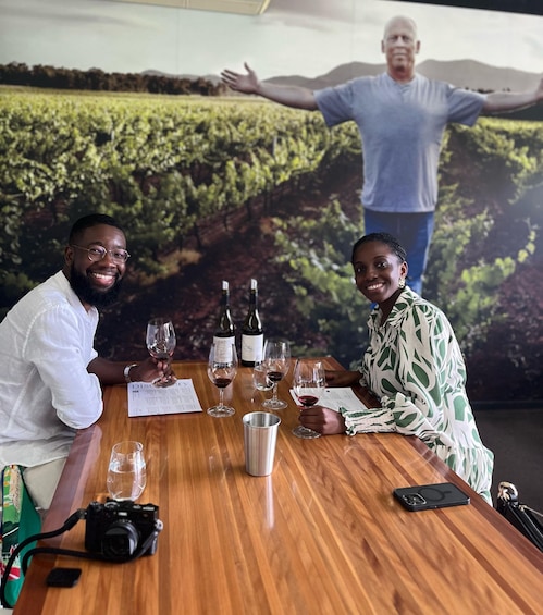 Two people sitting at a wooden table with wine glasses and bottles, a camera, and a smartphone. 1