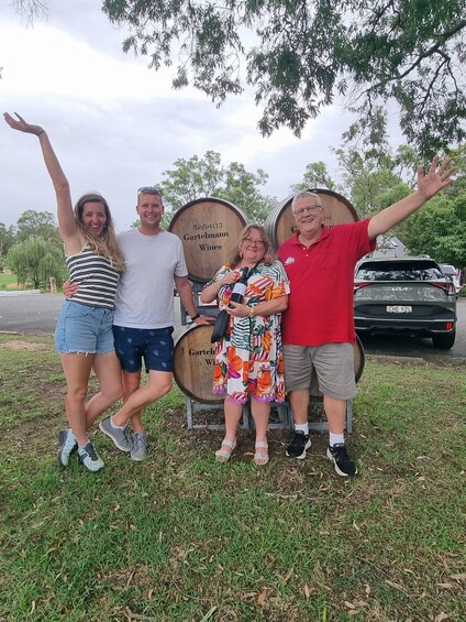 Four people standing in front of wooden barrels with text, a car, and trees in the background. 2