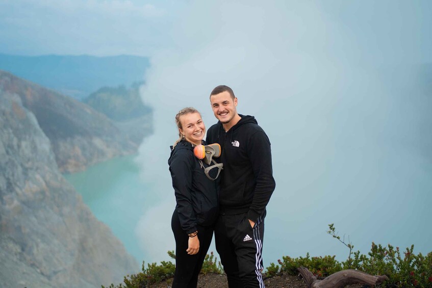 Two people standing on a cliff with a mountainous landscape and a body of water in the background. 1
