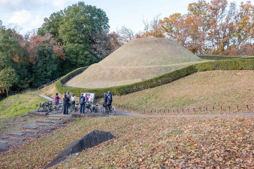A group of cyclists on a paved path, a large mound, and autumn trees. 2