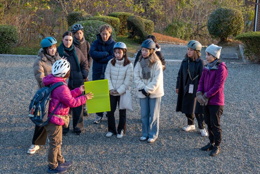 A group of people outdoors, some wearing helmets, gathered around a person holding a green folder. 3