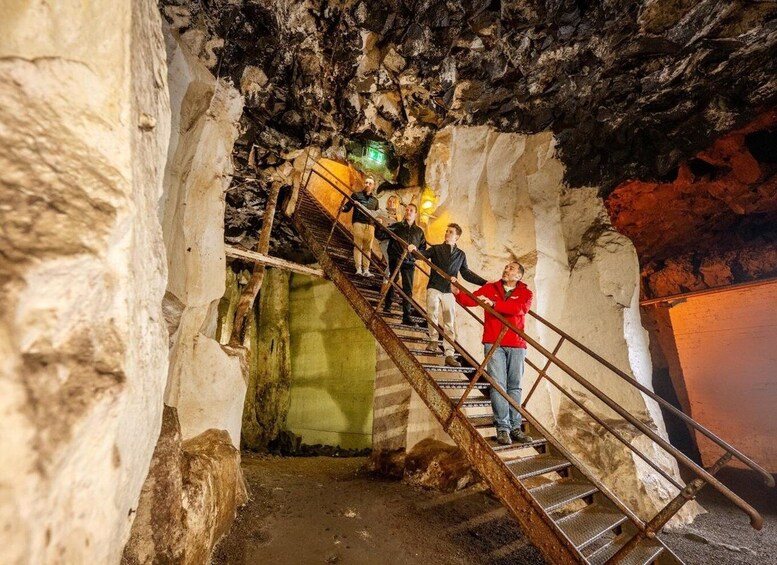 A group of people ascending a metal staircase in a cave with rocky walls and a green exit sign. 2