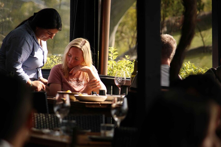 A restaurant with diners at a table, a wine glass, and greenery visible through the window. 2