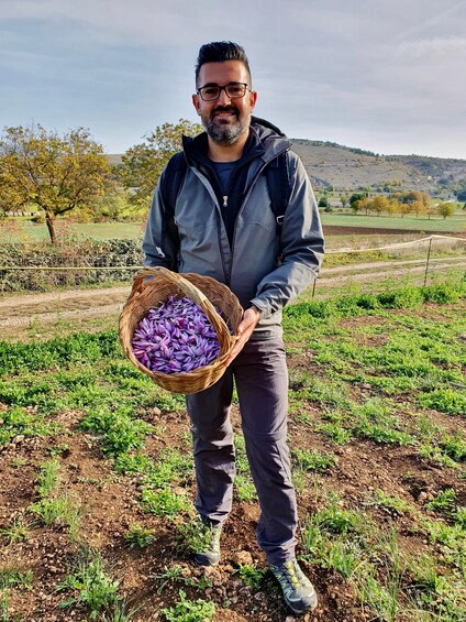 A person holding a basket of purple flowers in a field with a hilly landscape in the background. 2