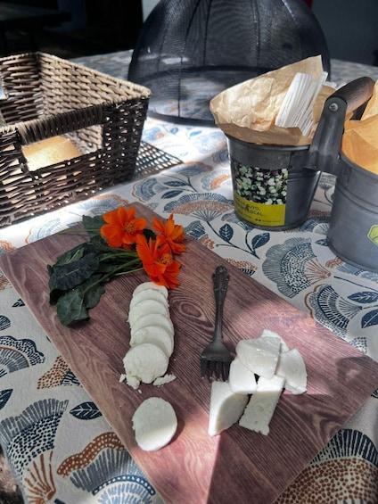 A wooden cutting board with sliced cheese, a fork, and a bouquet of flowers on a patterned tablecloth. 2