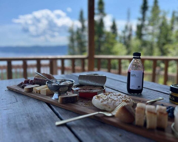 A wooden table with various cheeses, bread, and a bottle of wine on a deck with a scenic view of trees and a body of water. 4