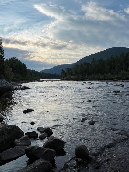 A river with rocks, surrounded by trees and hills under a cloudy sky. 2