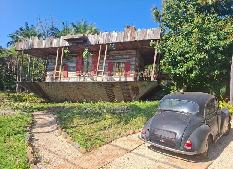 A vintage car parked in front of a wooden structure with a slanted roof, surrounded by greenery and a gravel path. 1