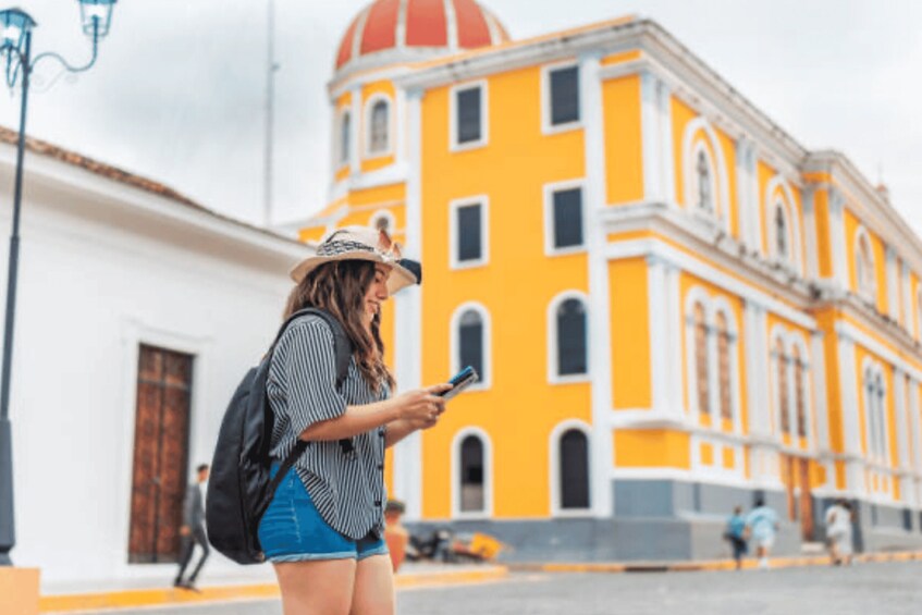 A person using a smartphone, standing in front of a yellow building with a dome and arched windows. 3