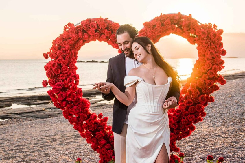 A couple standing in front of a heart-shaped floral arrangement on a beach at sunset. 3
