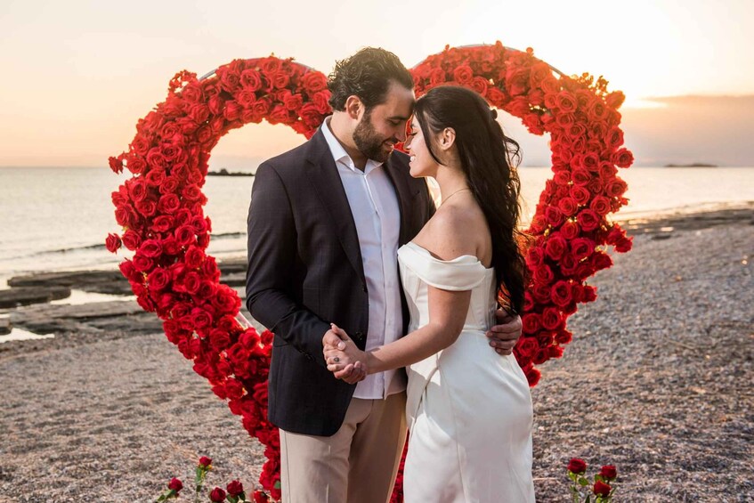 A couple standing in front of a heart-shaped arrangement of red roses on a beach with the ocean in the background. 5