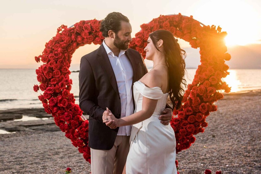 A couple holding hands in front of a heart-shaped arrangement of red roses on a beach at sunset. 2