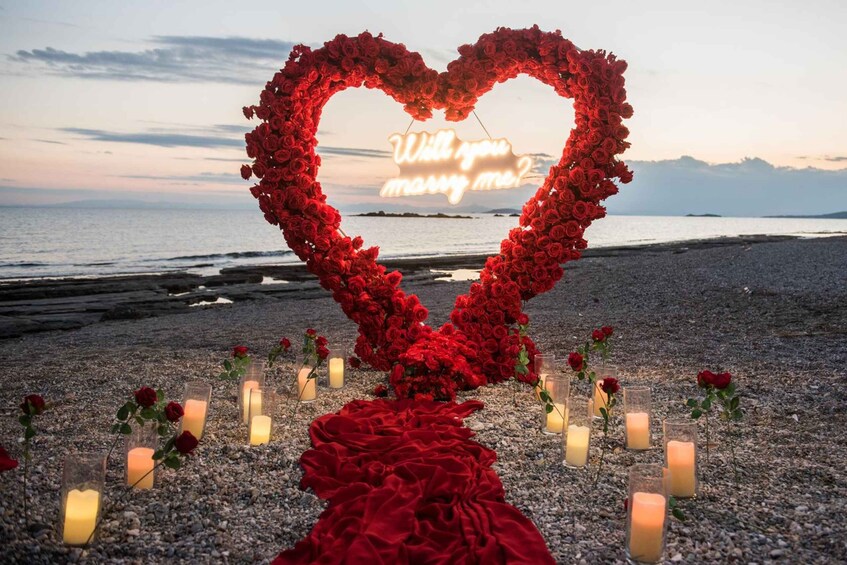 A heart-shaped arrangement of red roses on a pebbled beach with candles and a neon sign reading 'Will you marry me?' 4