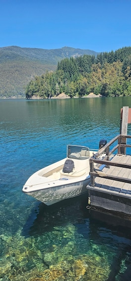 A boat docked at a pier with clear water and a forested hill in the background. 1