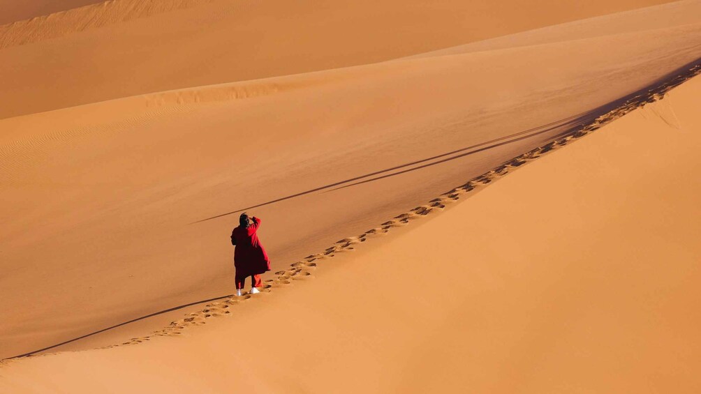 A person in a red coat walking on a sandy dune with footprints. 1