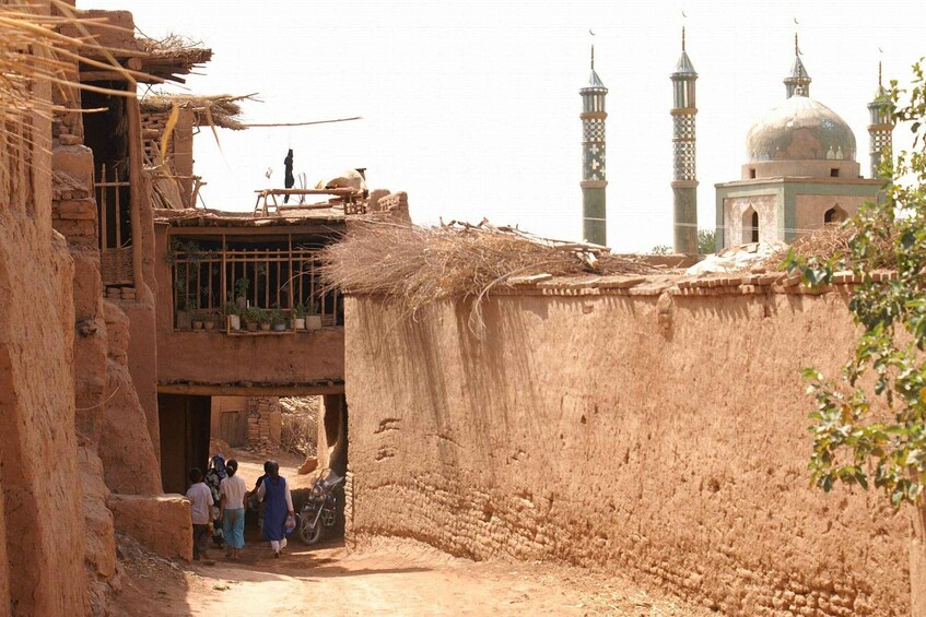 A rural village with mud-brick houses, a mosque with minarets, and a few people walking. 5