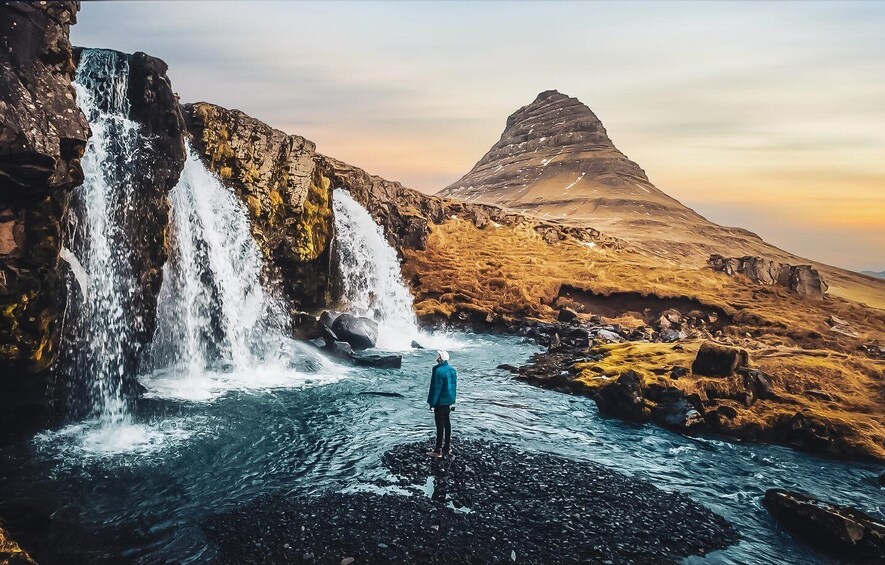 A person standing on a rocky outcrop looking at a waterfall and a mountain. 1