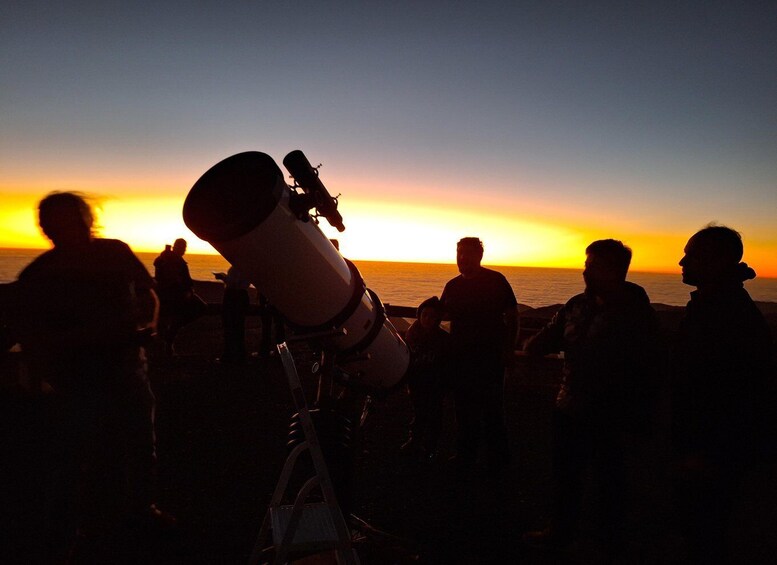 Ein Teleskop am Strand, vor einer Sonnenuntergangslandschaft mit silhouettierten Personen. 1