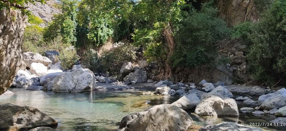 A clear stream with large rocks, surrounded by dense greenery and a rocky cliff. 1