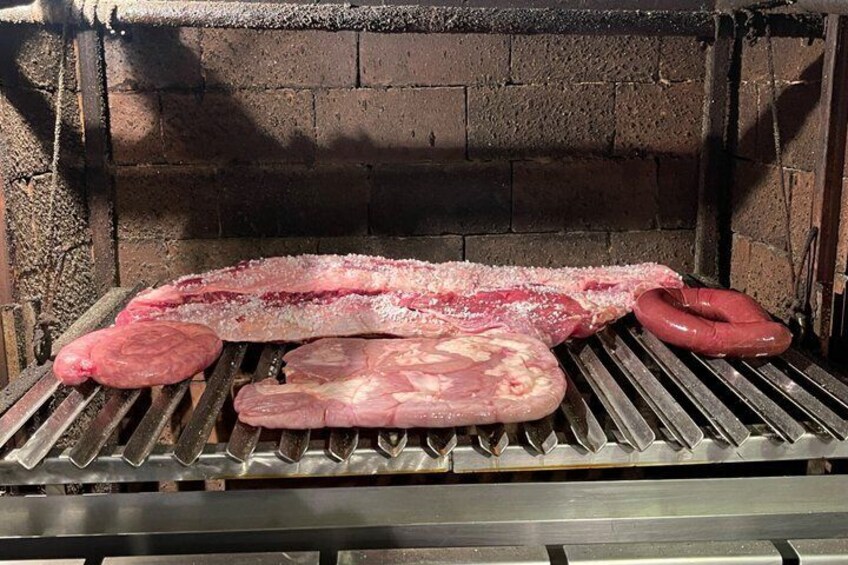 A meat grilling setup with sausages and a large piece of meat on a metal grate. 5