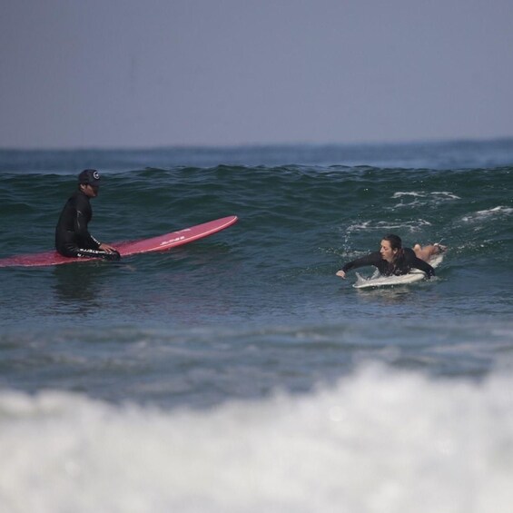 Two surfers on the water with waves, one standing on a red surfboard and the other lying on a white one. 5