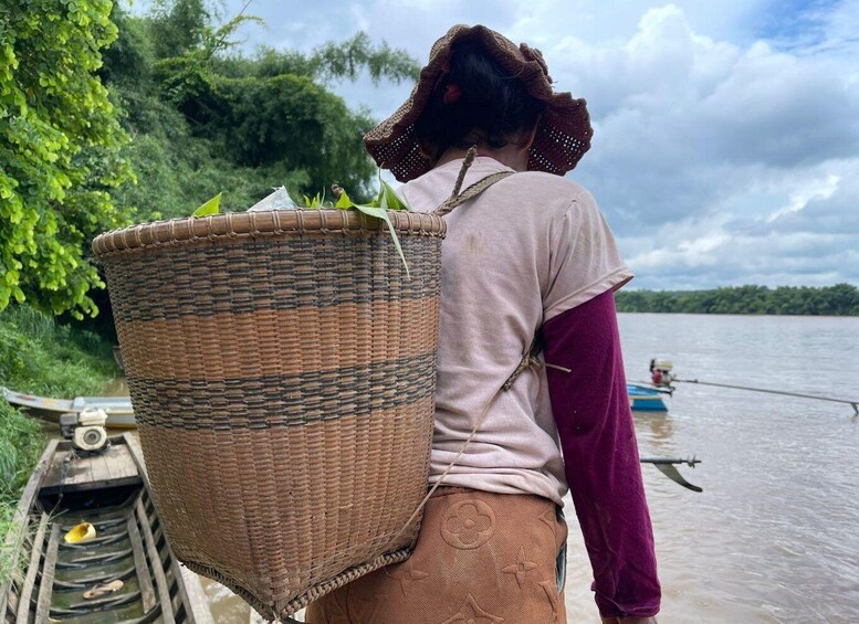 A person carrying a large woven basket on a boat, with a river and greenery in the background. 2