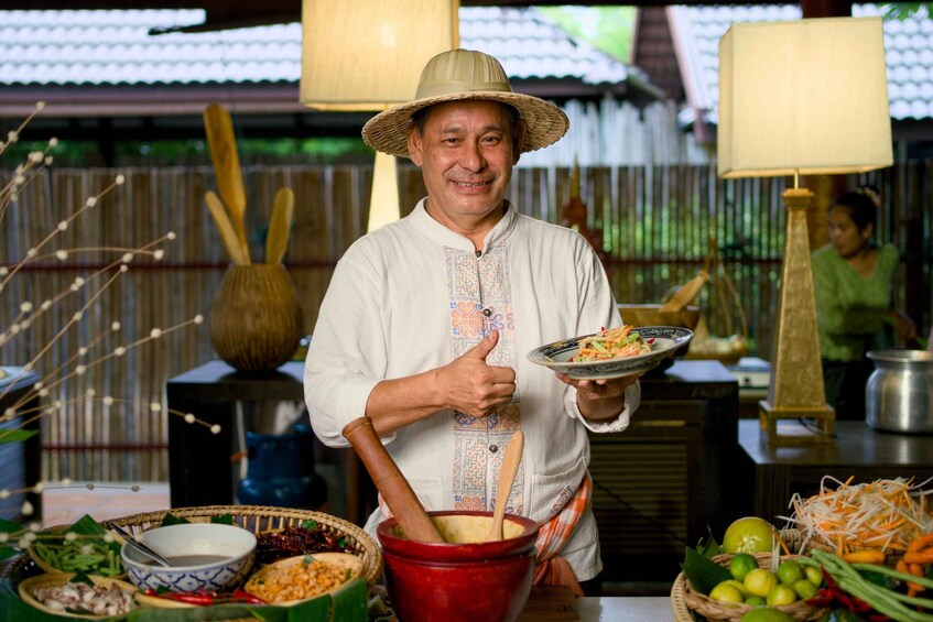 A person in a traditional hat holding a plate of food, with various dishes and ingredients on the table. 3