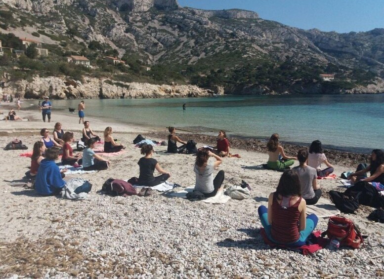 A group of people sitting on a pebble beach, facing the sea, with mountains in the background. 2
