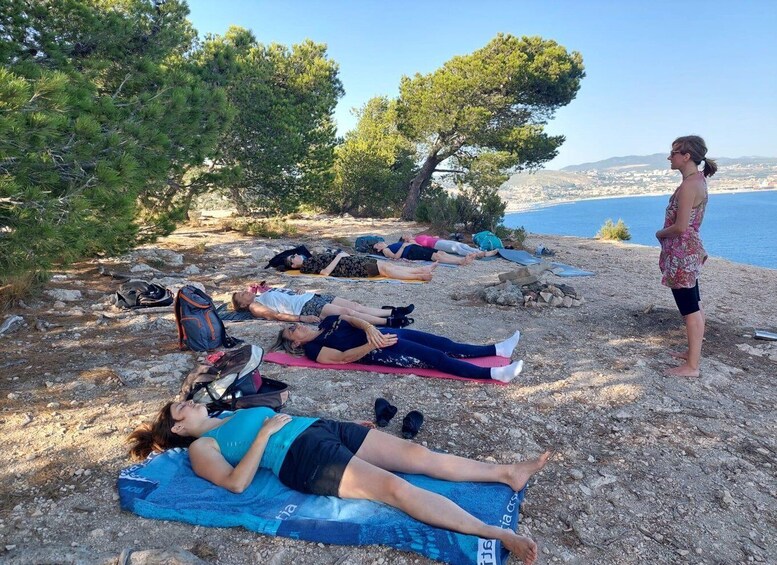 A group of people lying on mats outdoors, some on a rocky terrain and others near a body of water, with trees in the background. 5