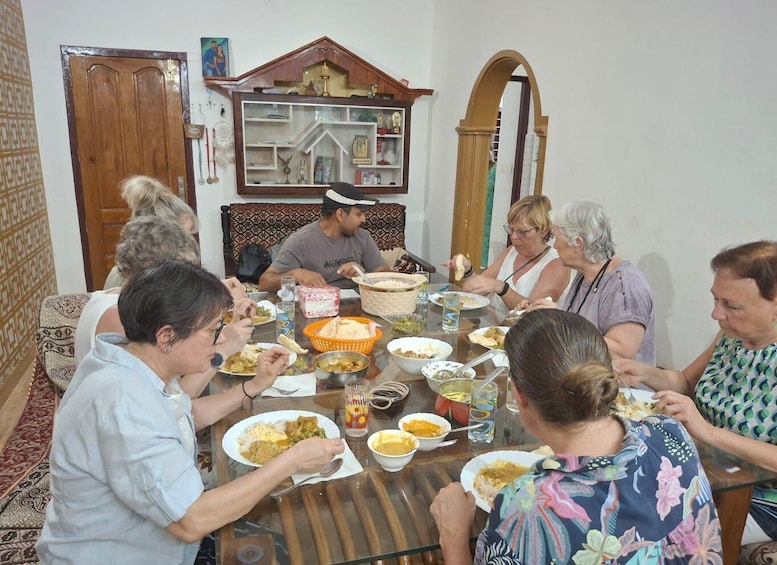 A group of people sitting around a table with various dishes, in a room with patterned walls and a wooden cabinet. 3