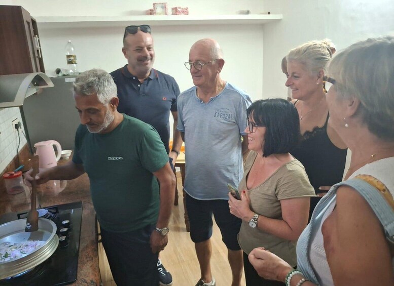A group of people in a kitchen, some standing and some seated, with a stove and cooking utensils visible. 5