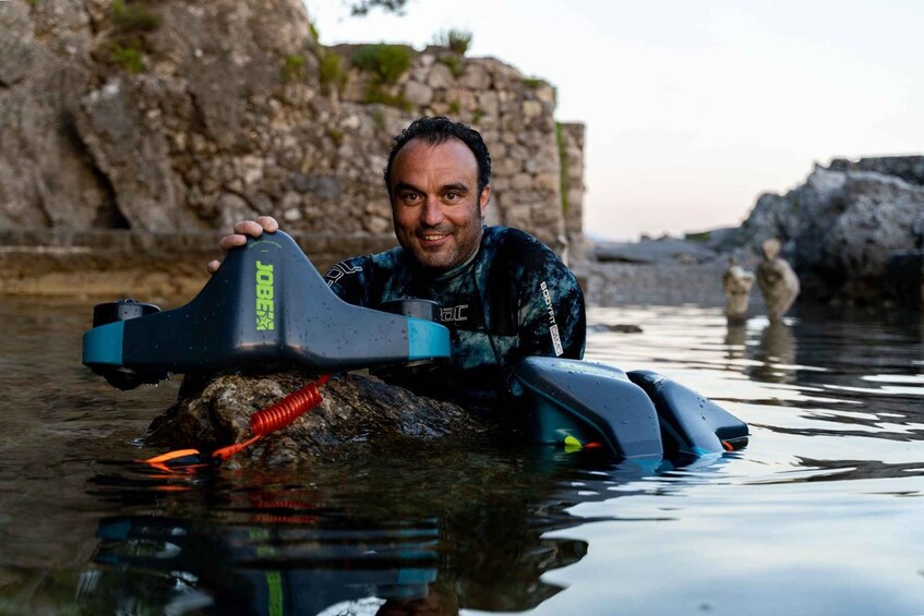 A person in a wetsuit holding a kayak paddle in a body of water with rocky terrain in the background. 1
