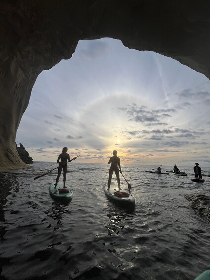People paddleboarding in a cave opening with the sun setting in the background. 3