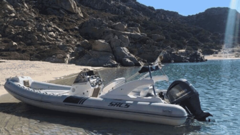 A white inflatable boat with a motor on a sandy beach with rocky cliffs in the background. 2