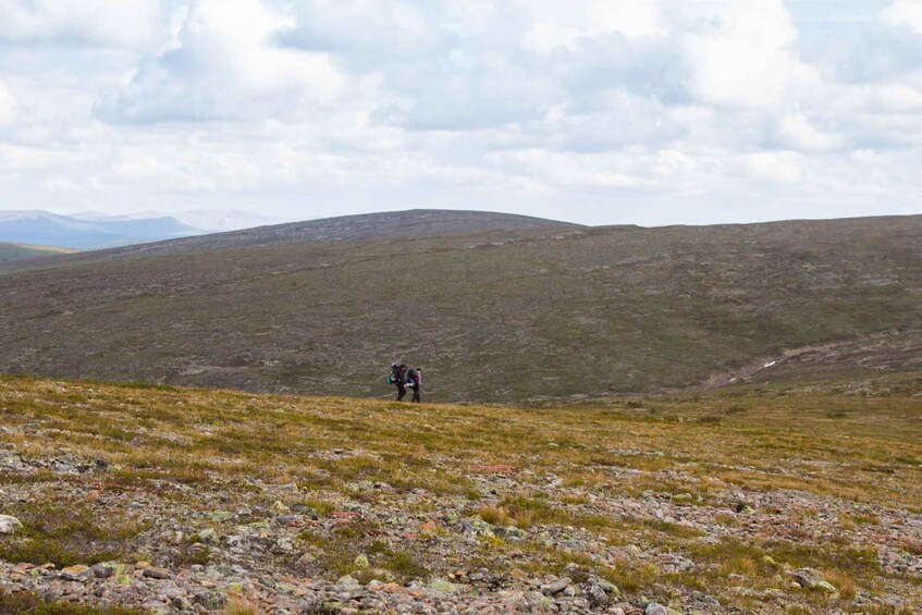 A hiker on a rocky, grassy hillside with distant mountains under a cloudy sky. 3