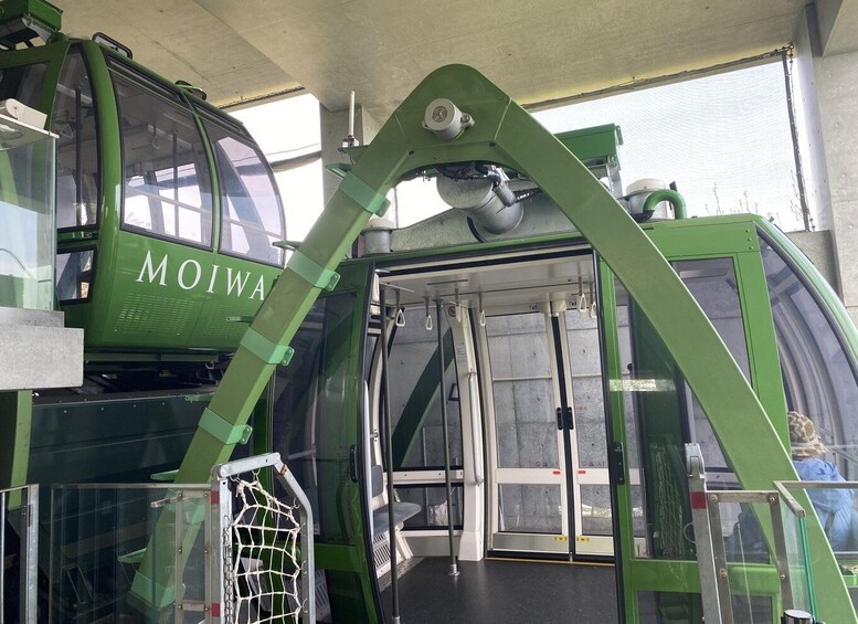 A green cable car with the word 'MOIWAR' on its side, parked at a station with a metal frame structure above it. 4