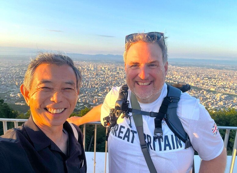 Two people standing on a viewing platform with a cityscape in the background. 1