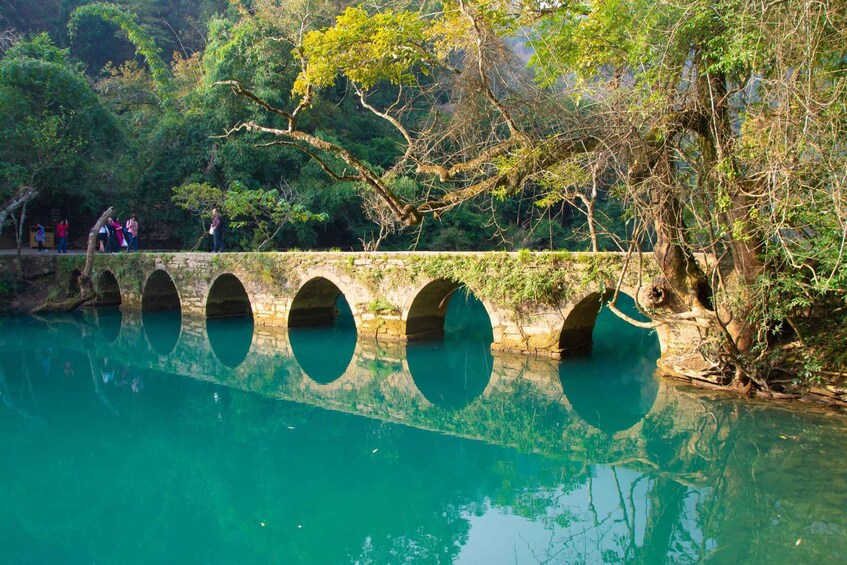 A stone bridge with multiple arches over a clear blue river, surrounded by lush greenery and trees. 1