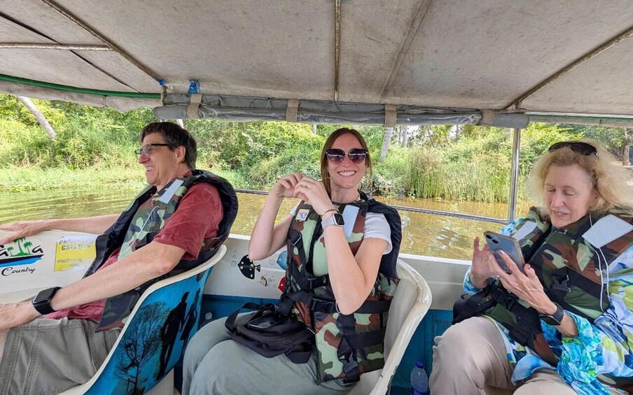 Three people on a boat wearing life jackets, with greenery in the background. 1