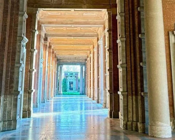 A long corridor with columns and a central walkway leading to a green area. 2