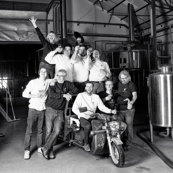 A group of people posing in a brewery setting with industrial equipment and a motorcycle. 2