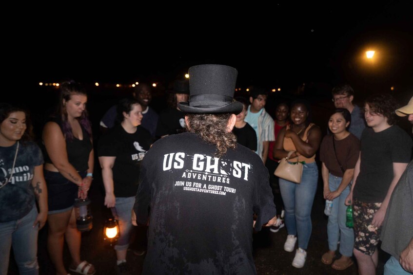 A group of people at a nighttime event, with one person in the foreground wearing a black shirt with 'US GHOST' text. 5
