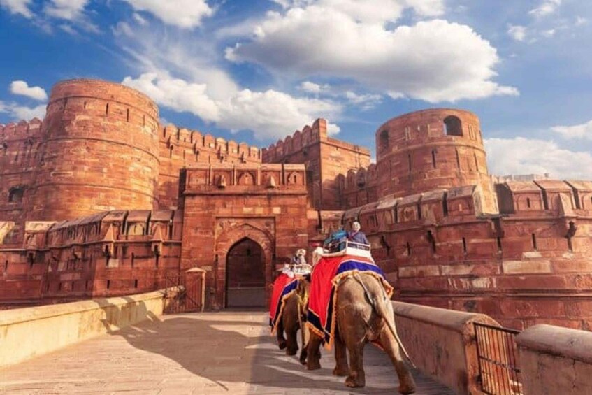 A historic fort with red brick walls and a large central gate, with elephants carrying people. 1