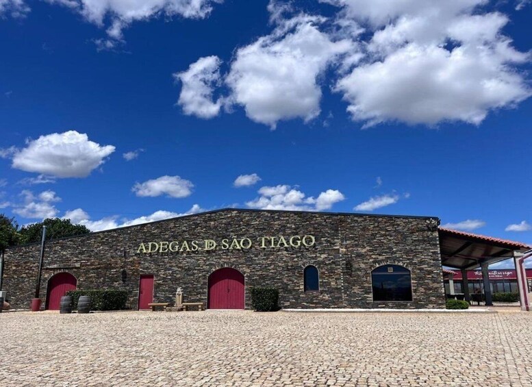 A stone building with the inscription 'ADEGAS DE SAO TIAGO' under a clear blue sky with scattered clouds. 1