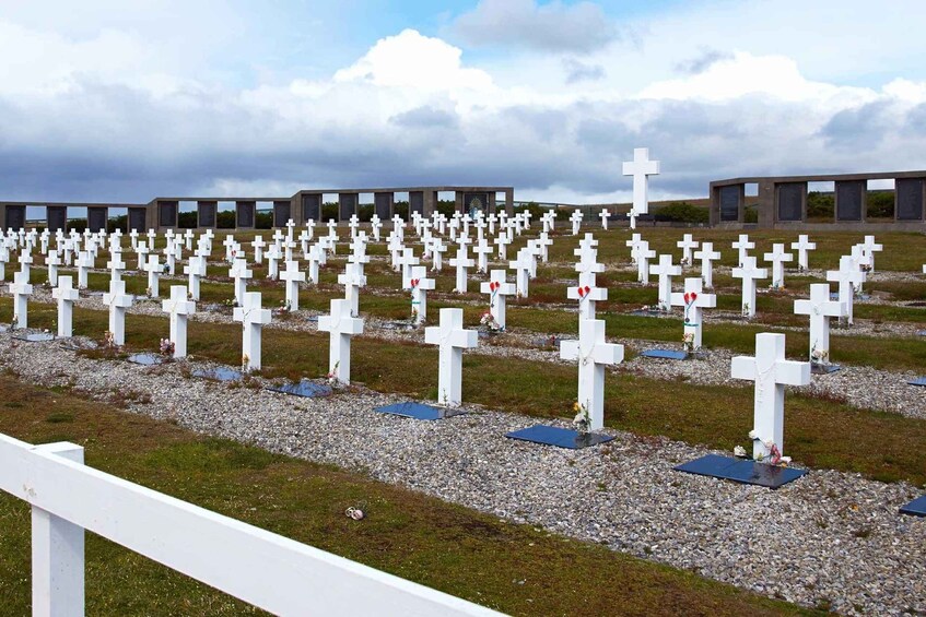 A cemetery with numerous white crosses, some adorned with flowers, under a cloudy sky. 3